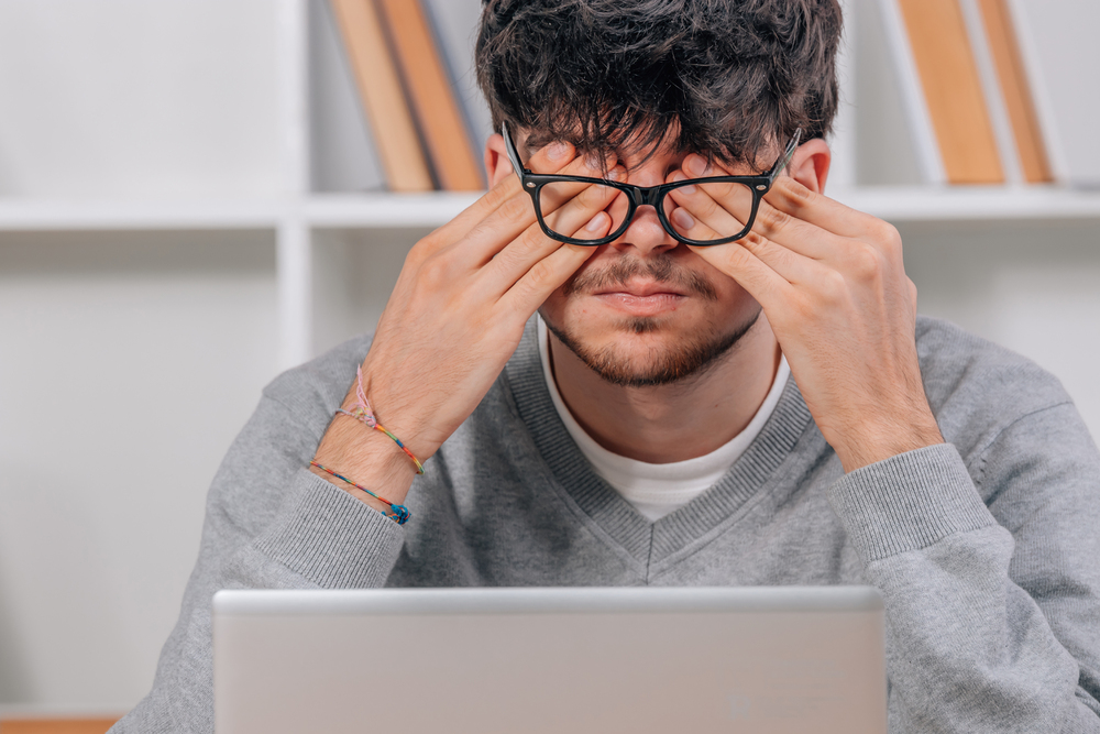 Junger Mann mit Computer, der sich vor Müdigkeit die Augen reibt als Symbolbild für den Ratgeber: Müde Augen