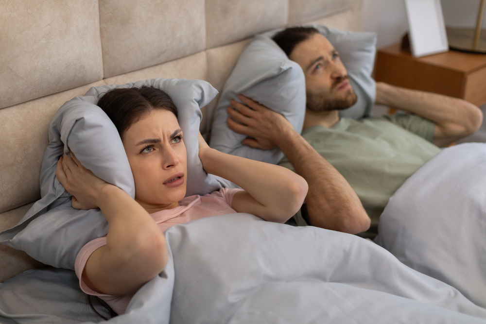 Junges Paar, das unter lauten Nachbarn leidet und sich die Ohren mit Kissen zuhält, während es nachts zu Hause im Bett liegt als Symbolbild für den Ratgeber: Junges Paar, das unter lauten Nachbarn leidet und sich die Ohren mit Kissen zuhält, während es nachts zu Hause im Bett liegt.