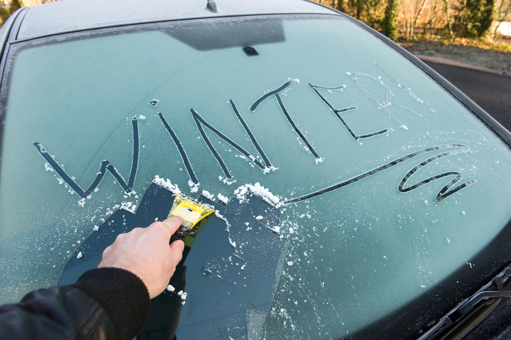 Ein EIskratzer in einer Hand, der dazu genutzt wird, eine Windschutzscheibem auf der das Wort Winter eingekratzt ist, von Frost zu befreien als Symbolbild für den Ratgeber: Scheibenenteiser selber machen