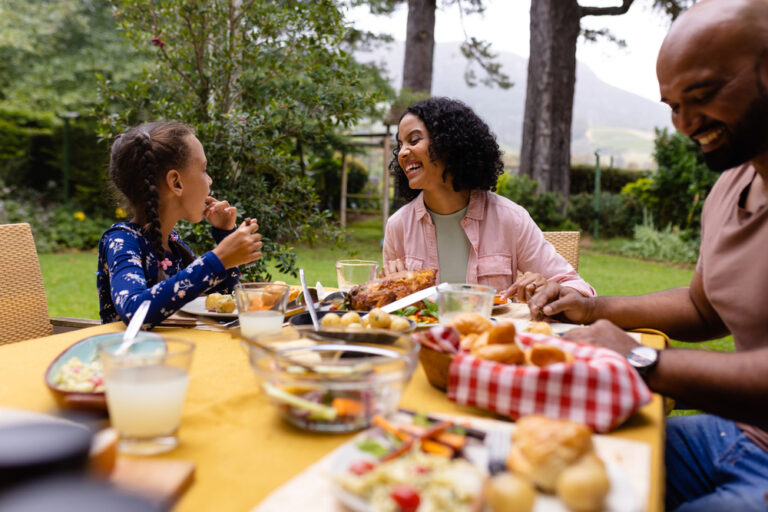 Eine Familie genießt ein gemeinsames Essen im Garten. Die Eltern lachen mit ihrem Kind, während sie sich angeregt unterhalten. Die warme Atmosphäre und das liebevolle Gespräch unterstreichen die Bedeutung wertschätzender Kommunikation in der Familie.