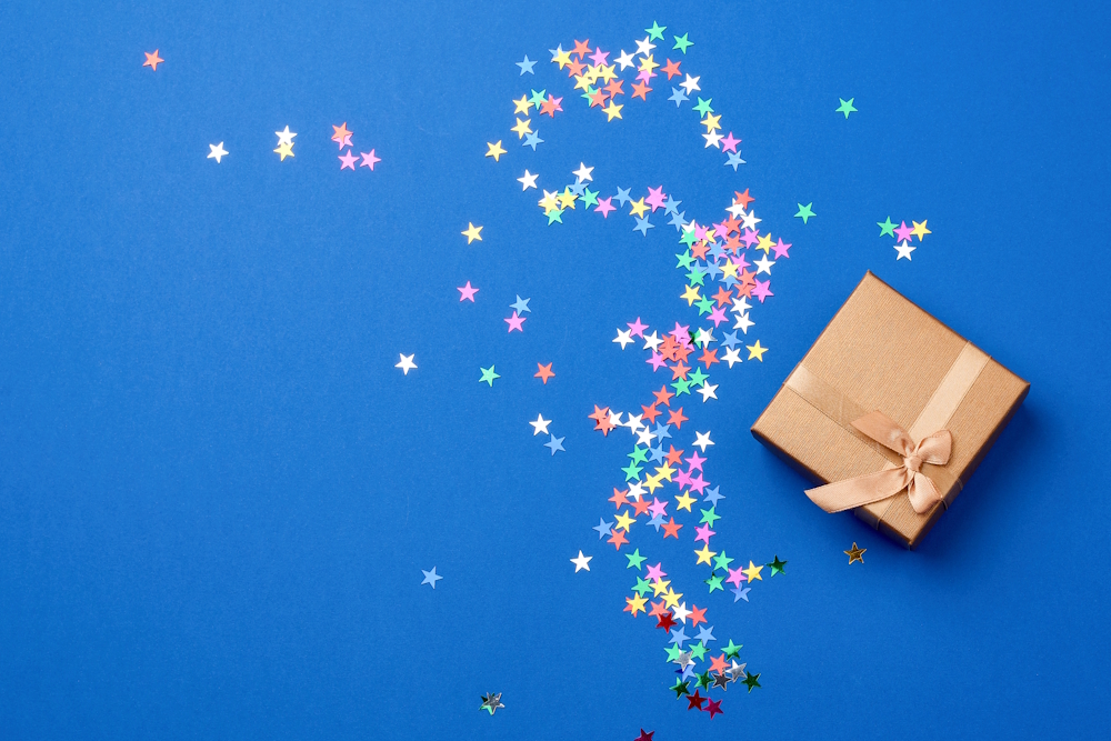 Geschenkbox mit Schleife, aus der bunte Sternkonfetti auf blauem Hintergrund wie symbolisierte Erlebnisse strömen.
