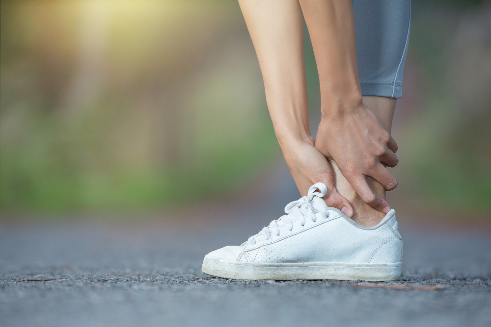 Frau hält sich schmerzhaft den umgeknickten Fuß mit weißem Sneaker auf Asphaltweg.