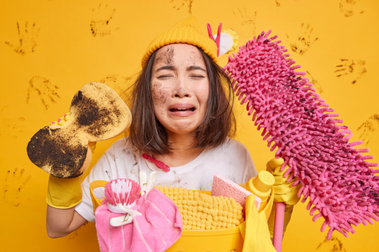 Frau mit gelber Mütze und Handschuhen, verschmutzt und verzweifelt beim Putzen mit Schwamm, Mop und Putzutensilien vor gelbem Hintergrund mit Handabdrücken.