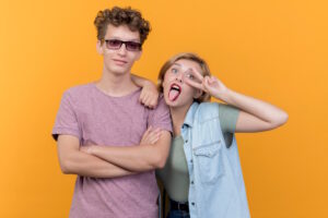 Junger Mann mit verschränkten Armen, junge Frau daneben macht Grimasse und zeigt Peace-Zeichen vor orangefarbenem Hintergrund.