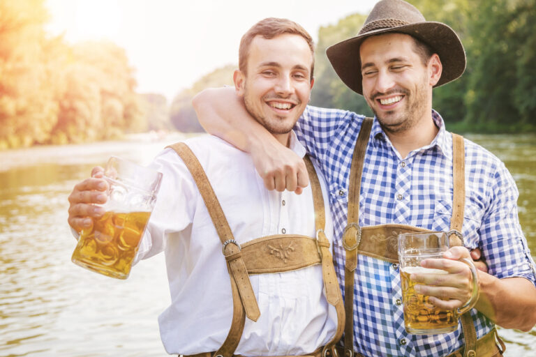 Zwei Freunde in bayerischer Tracht mit Bierkrügen am Fluss – Symbolbild zum Thema, woher der bayrische Ausdruck Spezi stammt.