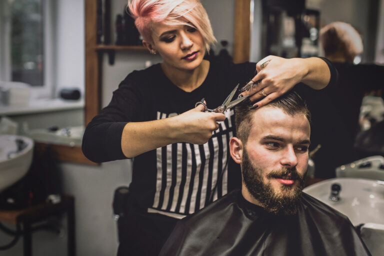 Friseurin mit kurzem, rosafarbenem Haar schneidet im Stehen einem Kunden die Haare (ein klassischer Stehberuf) in einem modernen Friseursalon.