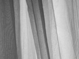 Feine, moderne Gardinen aus leichten, transparenten Stoffen in hellen Farben, sanft fallend in weichen Falten, vor einem großen Fenster. Die Stoffe variieren leicht in Struktur und Transparenz und schaffen eine helle, luftige Atmosphäre im Raum.