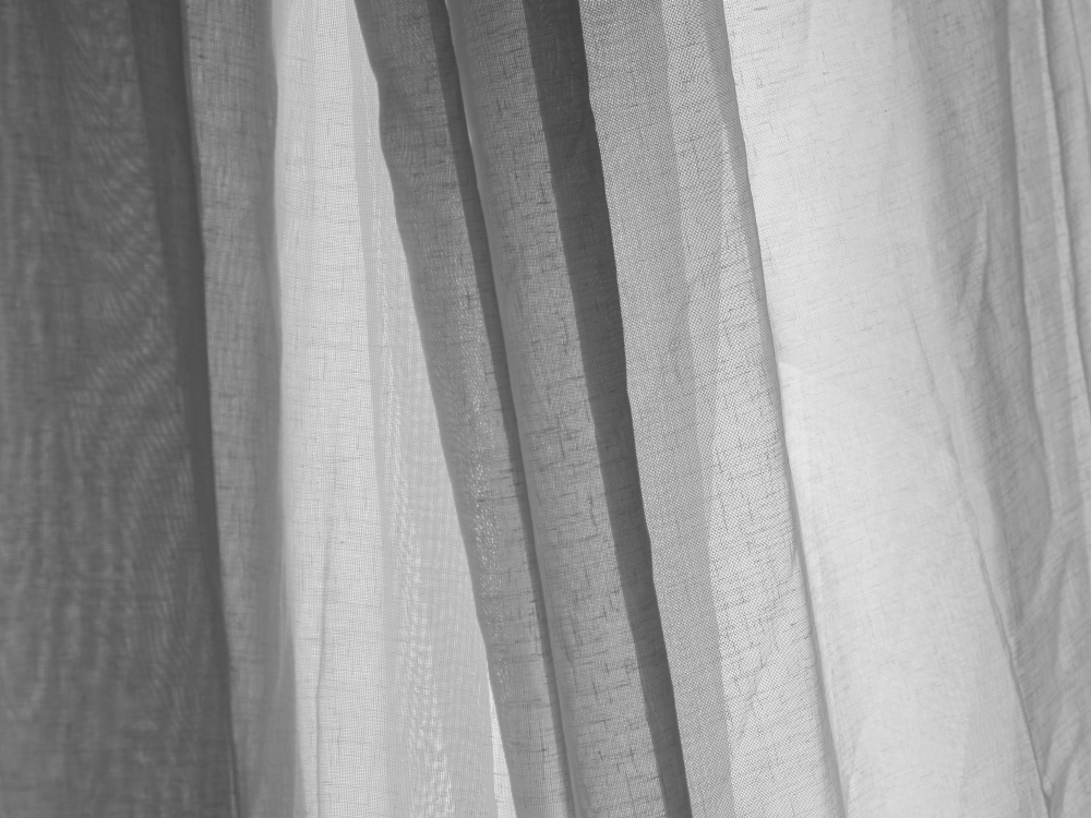 Feine, moderne Gardinen aus leichten, transparenten Stoffen in hellen Farben, sanft fallend in weichen Falten, vor einem großen Fenster. Die Stoffe variieren leicht in Struktur und Transparenz und schaffen eine helle, luftige Atmosphäre im Raum.