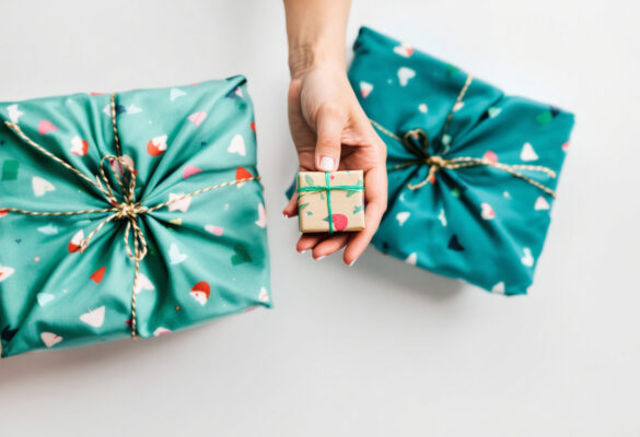 Eine Hand hält ein kleines, nachhaltig eingepacktes Geschenk vor zwei größeren Geschenken, die in wiederverwendbarem Stoff statt Geschenkpapier verpackt sind.