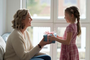 Ein Mädchen überreicht seiner lächelnden Mutter vor dem Fenster ein blaues Geschenk mit rosa Schleife, symbolisch für Geschenkideen zum Muttertag.