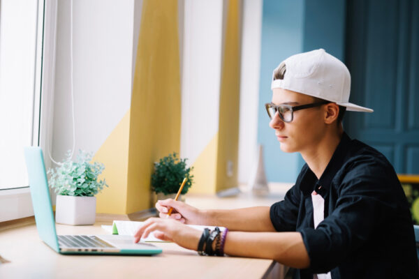 Ein junger Mann mit Brille und umgedrehter Baseballkappe sitzt konzentriert an einem Laptop und schreibt mit einem Stift Notizen. Das Bild symbolisiert das Thema 'Halbjahreszeugnis in der Bewerbung' und zeigt eine jugendliche Person, die sich auf den Bewerbungsprozess vorbereitet.