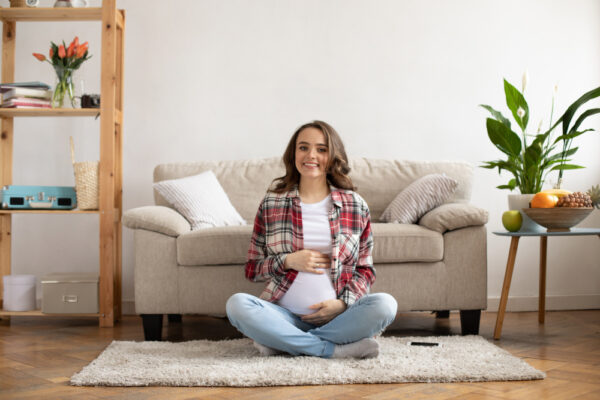 Eine schwangere Frau sitzt lächelnd im Schneidersitz auf einem Teppich in einem gemütlichen Wohnzimmer und hält sanft ihren Babybauch, symbolisch für die Frage: Hausgeburt – ja oder nein?