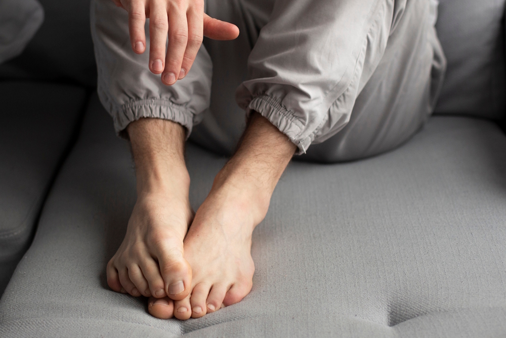 Eine Person in grauer Jogginghose sitzt auf einem grauen Sofa und legt ihre nackten Füße zusammen. Die Hand ist über den Füßen zu sehen. Der Fokus liegt auf den Zehen und Fußsohlen, passend zum Thema Hausmittel gegen Hühneraugen.