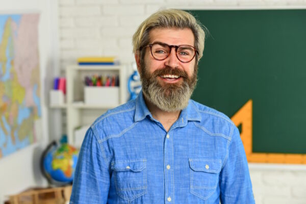 Ein lächelnder Lehrer mit Bart und Brille steht in einem Klassenzimmer, Symbolbild für Berufe, in denen man verbeamtet werden kann.