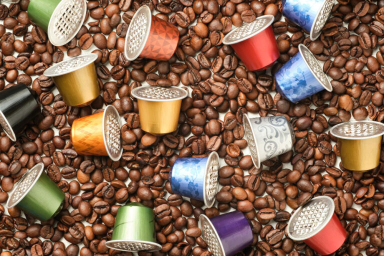 Mehrere bunte Kaffeekapseln verteilt auf einer großen Menge ganzer Kaffeebohnen. Kaffeekapseln oder Vollautomat – welche Variante ist die bessere Wahl?