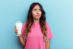 Junge Frau mit fragendem Blick auf hellblauem Hintergrund hält ein Glas Milch in der Hand und denkt nach