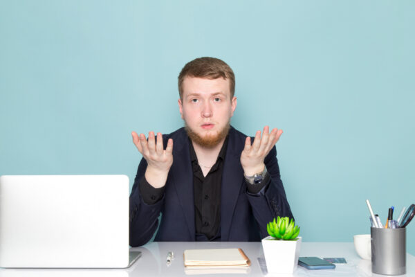 Ein junger Mann sitzt an einem Schreibtisch mit Laptop und Büromaterialien und hebt ratlos die Hände, als Symbol für das Warten auf eine Rückmeldung nach einer Bewerbung.