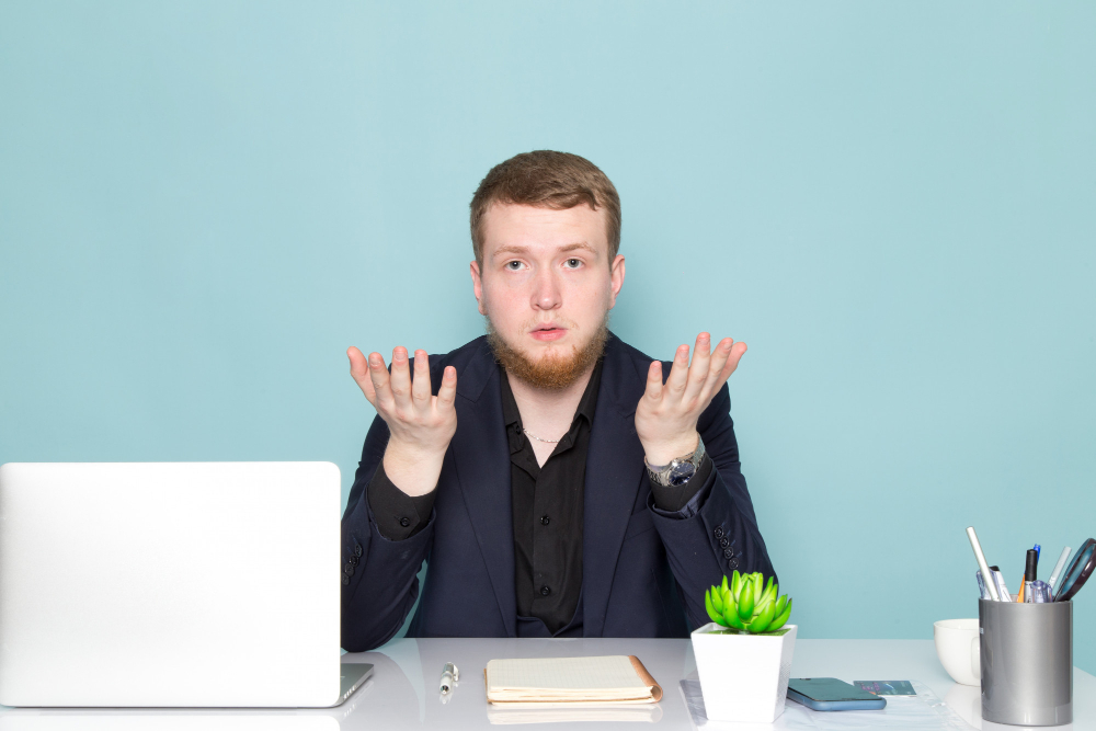 Ein junger Mann sitzt an einem Schreibtisch mit Laptop und Büromaterialien und hebt ratlos die Hände, als Symbol für das Warten auf eine Rückmeldung nach einer Bewerbung.