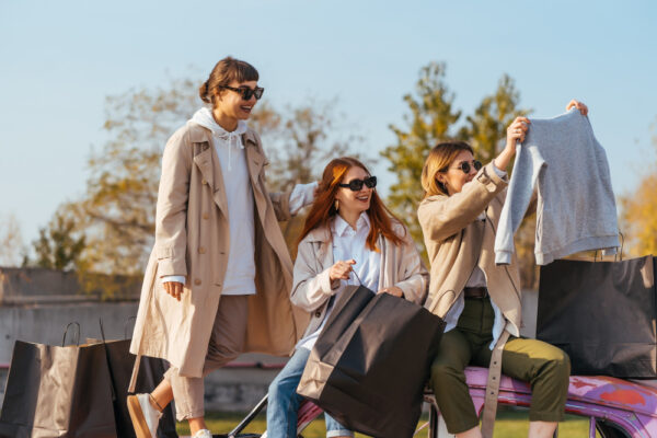 Drei Frauen mit Einkaufstaschen tauschen Kleidung im Freien und lachen gemeinsam.