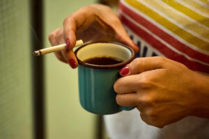 Eine Person mit roten Nägeln hält eine Tasse Kaffee in einer Hand und eine brennende Zigarette in der anderen – eine symbolische Darstellung von Koffein und Nikotin im Alltag.
