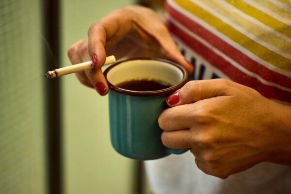 Eine Person mit roten Nägeln hält eine Tasse Kaffee in einer Hand und eine brennende Zigarette in der anderen – eine symbolische Darstellung von Koffein und Nikotin im Alltag.