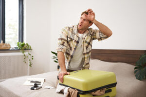 Mann mit erschöpftem Gesichtsausdruck sitzt krank auf dem Bett und hält Stirn, neben ihm ein offener Koffer.