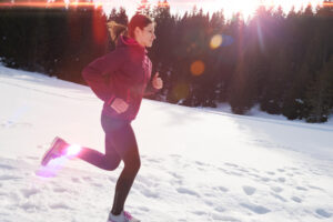 Frau joggt bei Sonnenschein durch verschneite Winterlandschaft vor dunklem Tannenwald.