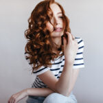 Frau mit langen, roten Locken in gestreiftem T-Shirt vor hellem Hintergrund.