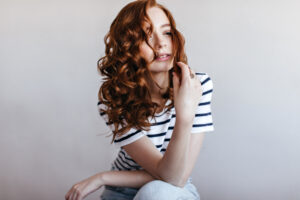 Frau mit langen, roten Locken in gestreiftem T-Shirt vor hellem Hintergrund.