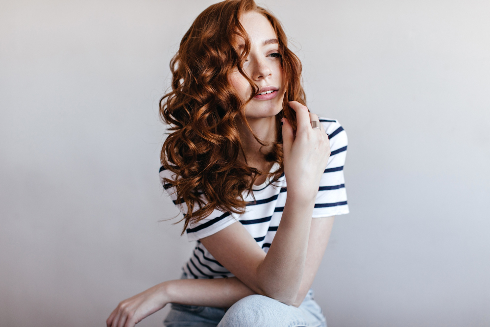 Frau mit langen, roten Locken in gestreiftem T-Shirt vor hellem Hintergrund.