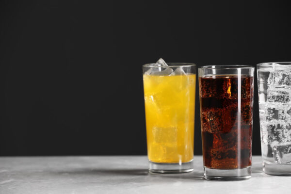 Fanta, Cola und Zitronenlimonade in drei Gläsern mit Eiswürfeln, vor einem dunklen Hintergrund aufgestellt. Das Bild symbolisiert das Thema 'Machen Softdrinks die Zähne kaputt?' und verweist auf die potenziell schädliche Wirkung von zuckerhaltigen und säurehaltigen Getränken auf die Zahngesundheit.