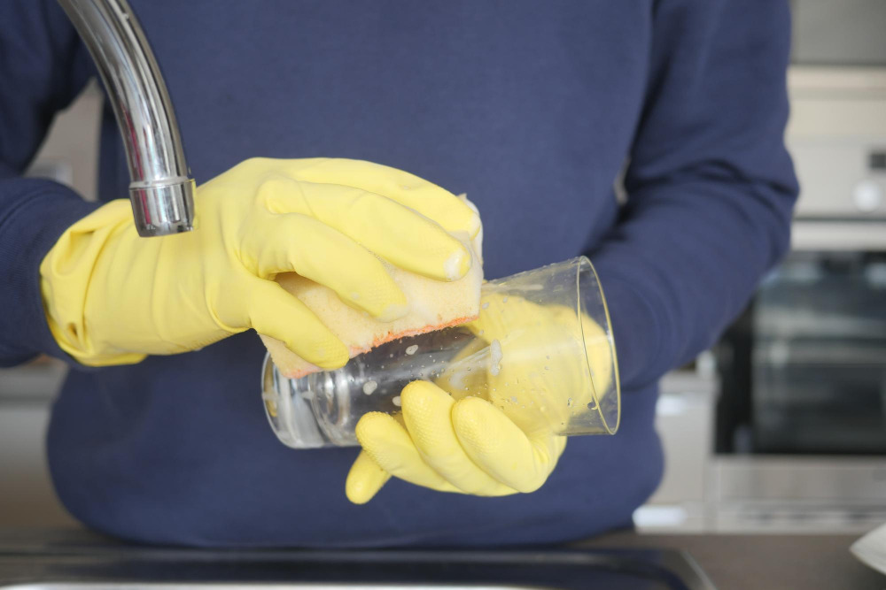 Mann wäscht mit gelben Putzhandschuhen Rückstände von einem Trinkglas ab