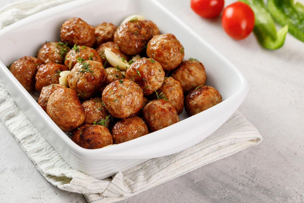 Mini-Frikadellen in einer weißen Auflaufform, garniert mit Kräutern und Knoblauch, daneben Tomaten und grüne Paprika.