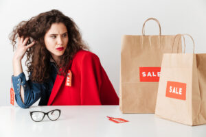 Frau mit nachdenklichem Blick sitzt vor Einkaufstüten mit „Sale“-Aufdruck und wirkt unzufrieden mit ihrem Kaufverhalten.
