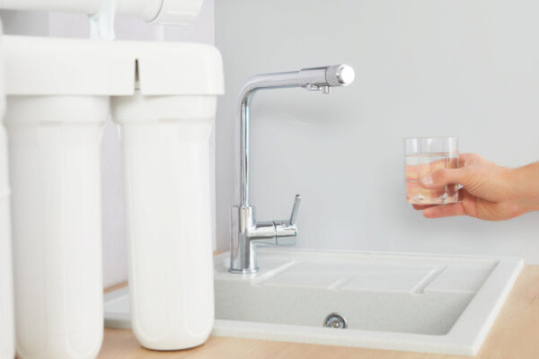 Osmose Wasserfilter im Vorderung und Hand mit Glas, die im Hintergrund Wasser aus dem Wasserhahn holen möchte.
