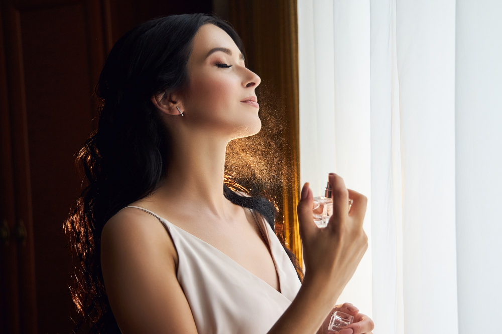 Frau mit dunklen Haaren sprüht Parfum auf ihren Hals vor einem hellen Fenster.