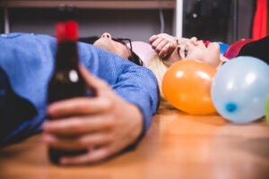 Zwei Personen liegen nach einer exzessiven Party auf dem Boden: Der Mann (im blauen Hemd) liegt erschöpft und mit einer Flasche in der Hand im Vordergrund, während die Frau im Hintergrund, umgeben von Ballons, sichtbar erschöpft vom Umgang mit seinem Rausch ihre Hand an die Stirn legt.