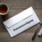 Weißer Briefumschlag mit der Aufschrift „Patientenverfügung“ auf einem Holztisch, daneben ein Kugelschreiber und eine Tasse Tee.