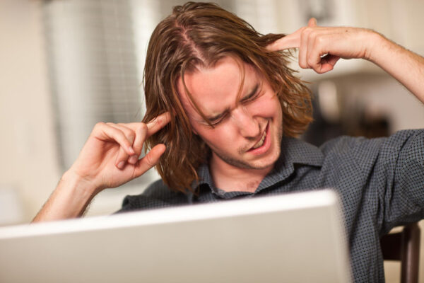 Ein junger Mann sitzt vor einem Laptop und hält sich mit schmerzverzerrtem Gesicht die Finger in die Ohren – eine Darstellung des Themas Piepen im Ohr und möglichen Symptomen von Tinnitus.