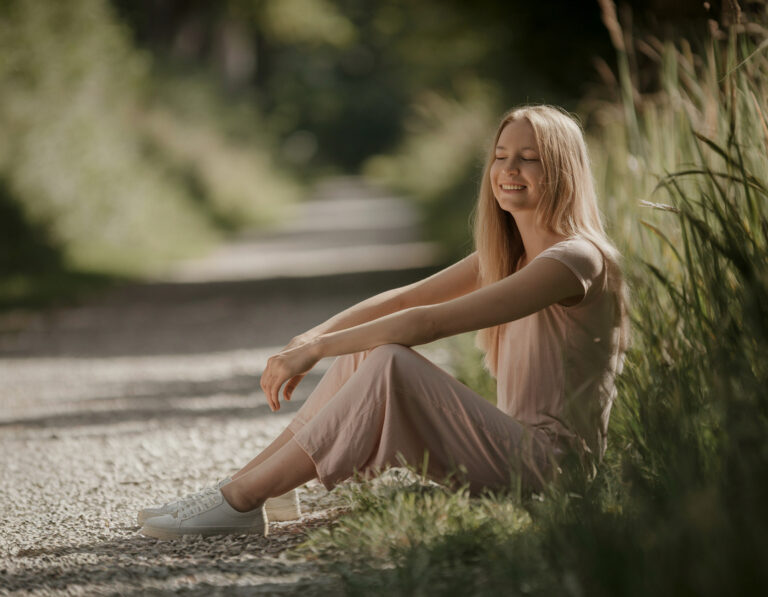 Junge Frau mit langen blonden Haaren sitzt entspannt auf einem Naturweg, die Augen geschlossen und lächelnd, umgeben von Grün – Symbol für Gelassenheit und Frugalismus.
