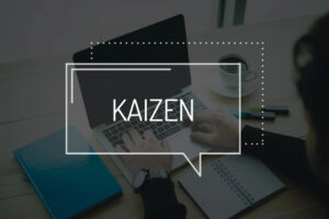 Laptop-Arbeitsplatz mit Notizbuch, Stiften und Kaffee, überlagert mit dem Wort 'KAIZEN'.