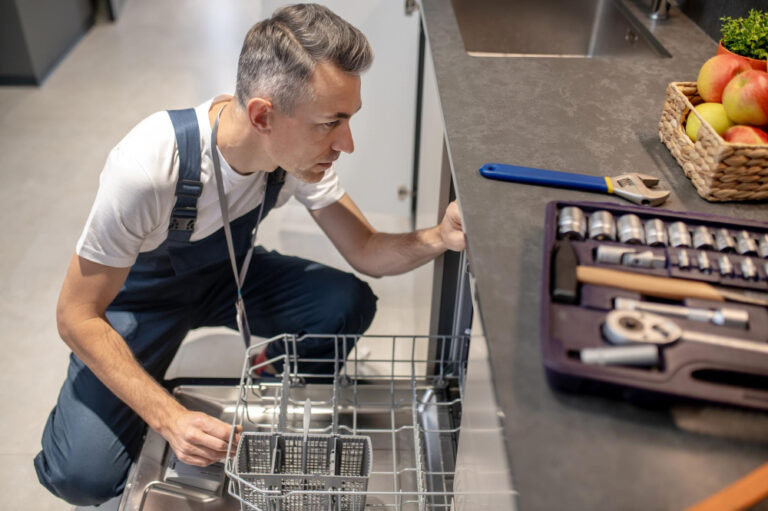 Handwerker inspiziert eine Spülmaschine mit ausgebreitetem Werkzeug auf der Arbeitsplatte.