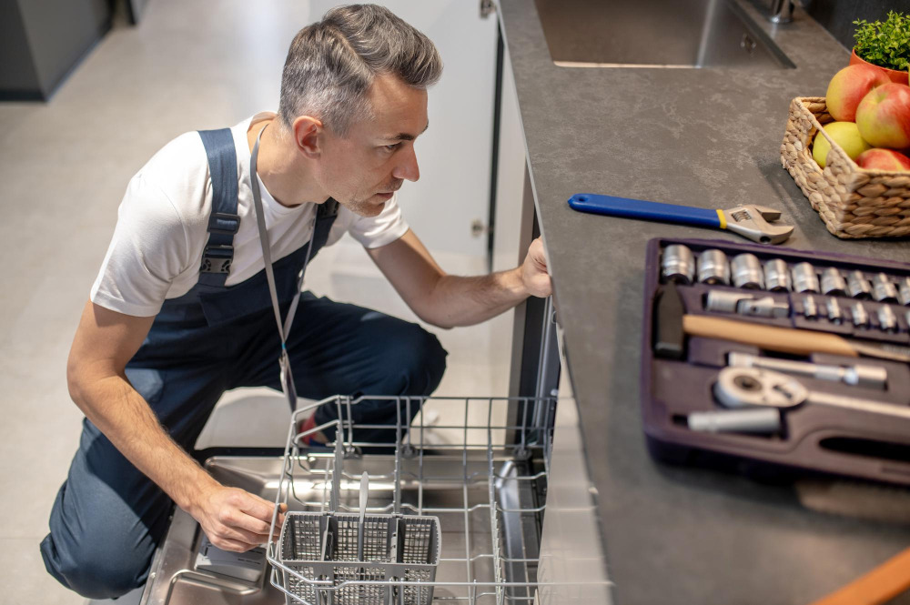 Handwerker inspiziert eine Spülmaschine mit ausgebreitetem Werkzeug auf der Arbeitsplatte.