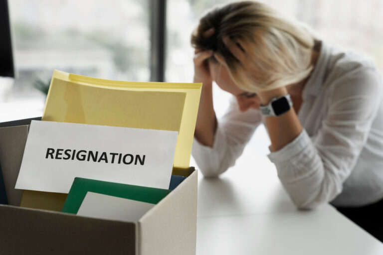 Frau sitzt mit gesenktem Kopf am Schreibtisch, im Vordergrund Karton mit Akten und Schild „Resignation“.