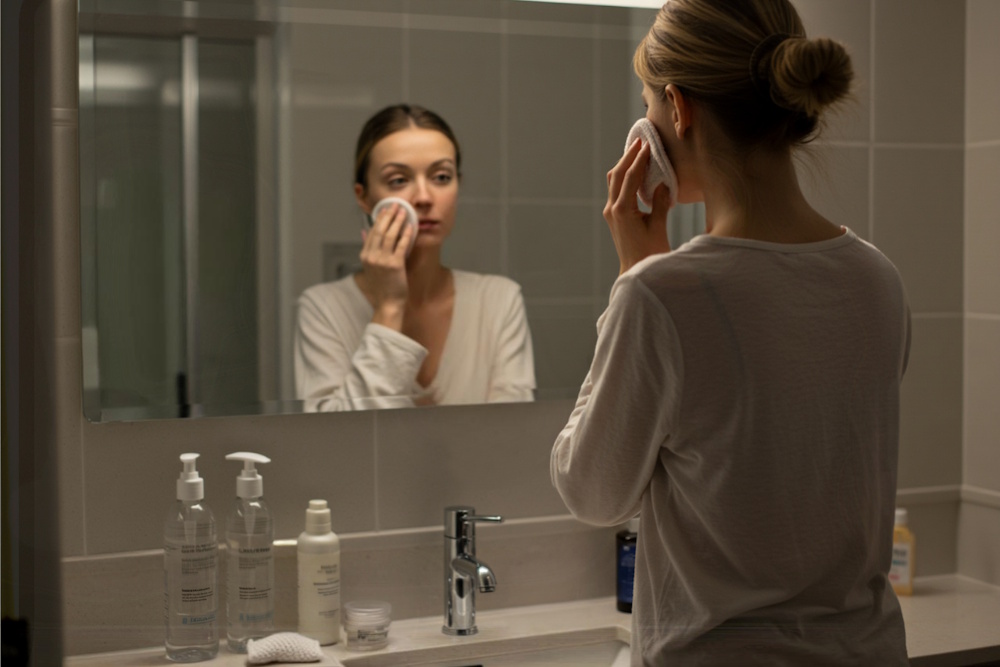 Junge Frau von hinten im Badezimmer, während sie ihr Gesicht mit einem Mikrofaserpad abschminkt – betont eine ruhige Routine und richtiges Abschminken.