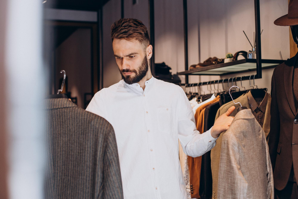 Mann in weißem Hemd betrachtet zwei Sakkos in einem Secondhand-Modegeschäft.