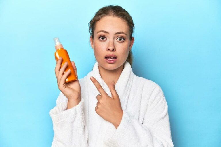 Junge Frau im weißen Bademantel hält eine Sonnencreme in ihrer rechten Hand und zeigt mit dem Finger ihrer linken Hand darauf als Symbolbild für den Ratgeber: Sonnencreme-Flecken entfernen