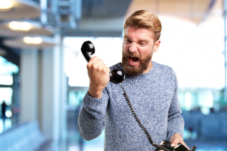 Wütender Mann mit Bart schreit in den Hörer eines schwarzen Telefons.