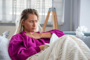 Frau hält sich den Hals und sieht krank aus, Taschentücher liegen daneben