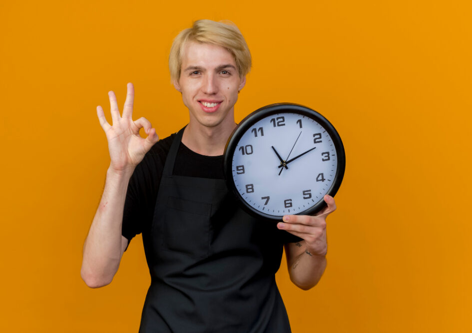 Ein junger Mann im schwarzen T-Shirt hält eine große Uhr und zeigt mit der anderen Hand das OK-Zeichen vor einem gelben Hintergrund, symbolisiert das Thema Minuten in Stunden umrechnen.